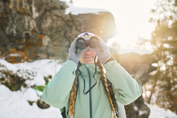 Caucasian young adult woman with long braids standing outdoors in snowy landscape using binoculars for observing distant scenery, sunlight shining behind rocky background