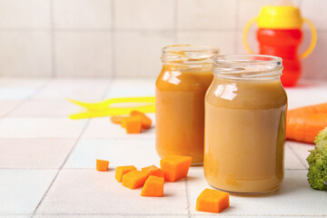 Glass jars of healthy baby food and vegetables on light tile background