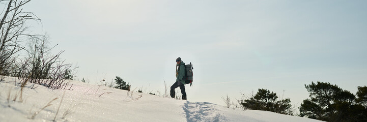 Caucasian middle aged man hiking through snowy landscape carrying large backpack, walking along snow covered trail with trees and dry grass in background under clear sky