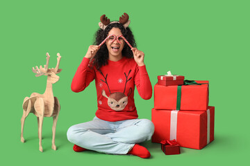 Beautiful young African-American woman in reindeer horns with candy canes and Christmas gift boxes sitting on green background