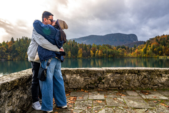 A newly engaged couple kisses and embraces by Lake Bled, Slovenia, surrounded by autumn trees and calm waters, capturing a pure moment of love, joy, and romantic connection.