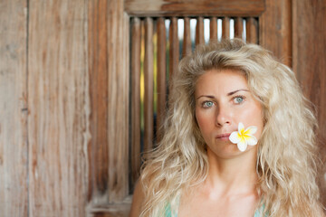 Young blonde woman with frangipani flower on open-air veranda of wooden bungalow in tropical garden. Girl relaxing in luxury villa on family summer vacation. Beach travel, spa, natural lifestyle.