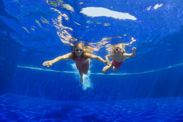 Happy family - mother, baby girl in goggles swim, dive in pool with fun - jump deep down underwater. Healthy lifestyle, people water sport activity, swimming lessons on holidays with kids