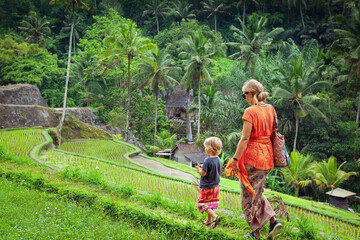 Mother and child walk along flooded lush rice terraces in Bali, wearing traditional sarongs, enjoying tropical nature and travel moments during a cultural family journey in Gunung Kawi in Indonesia.