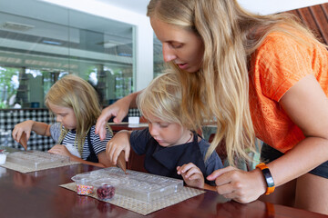 Kids and their mother participate in a chocolate making workshop in Bali, decorating sweets with toppings. A fun and educational family activity during tropical holidays.
