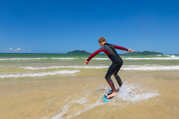 Young boy skimboarding in shallow water on a sunny beach day. Wearing wetsuit, balancing on board with determination and focus. Perfect image for themes of childhood adventure, active lifestyle, water