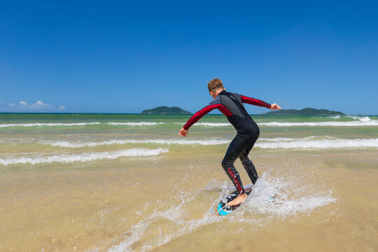 Young boy skimboarding in shallow water on a sunny beach day. Wearing wetsuit, balancing on board with determination and focus. Perfect image for themes of childhood adventure, active lifestyle, water