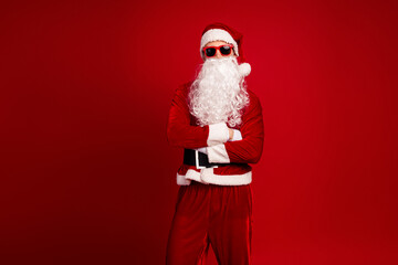Santa in a red suit with white beard belt with crossed arms wearing sunglass against a bold red background for christmas season promotion