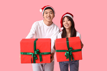 Beautiful young Asian couple in Santa hats with gift boxes on pink background. Christmas celebration