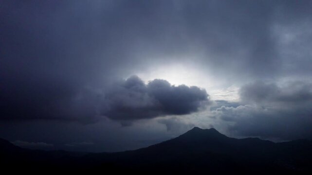 A dramatic, moody shot of dark storm clouds passing over a mountain silhouette. Bright light breaks through, creating an ominous and epic landscape.
