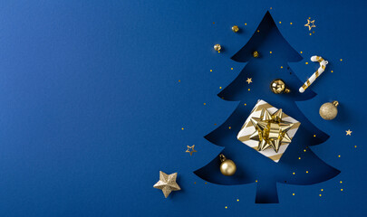 A blue backdrop hosts a dark blue Christmas tree cutout, gold gift box with bow, ornaments, and...
