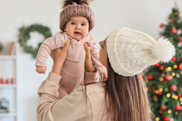 Happy mother with her little baby in warm hats at home on Christmas eve