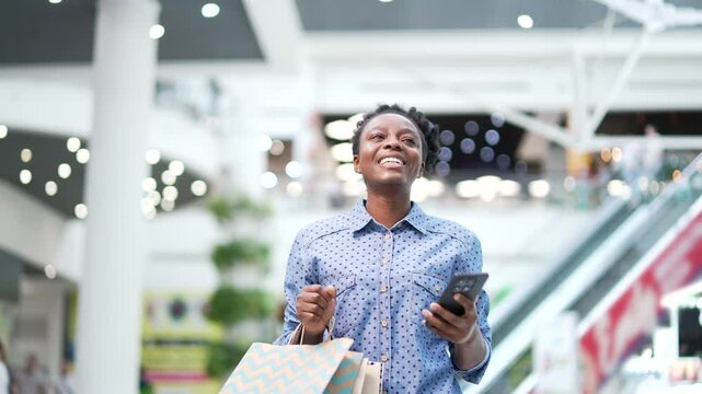 Young African American female shopper walking in shopping center mall. Smiling black woman with gift packages bag holding phone smartphone in hand, satisfied with sale discount day or black friday