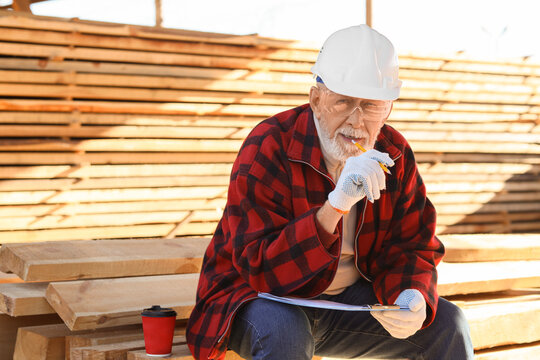 Happy mature male carpenter in hardhat with clipboard and cup of coffee sitting at sawmill - Powered by Adobe