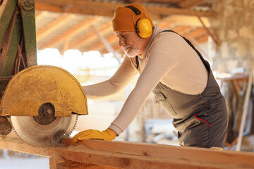 Mature male carpenter sawing wooden plank at sawmill
