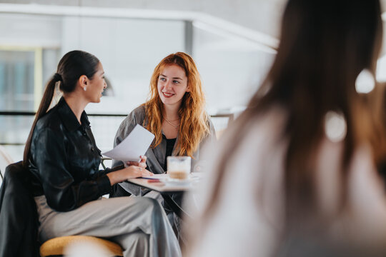 Two young women share a friendly discussion over papers and coffee in a bright cafe, suggesting teamwork, networking, and casual business meetings in a modern workspace.