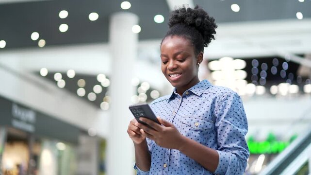 Young smiling african american woman shopper using mobile phone standing in shopping center mall. Happy joyful black woman customer browsing web, chatting online, typing using smartphone in store