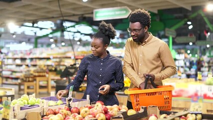 African American family couple customers buyers chooses fruits in a grocery store, market or supermarket. Man and woman makes daily purchases buy in retail shop store with basket select pick products