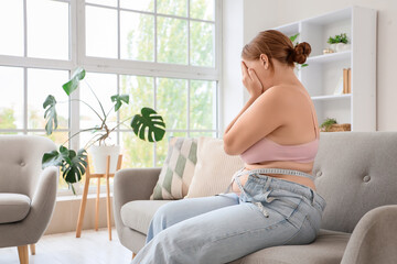 Upset overweight woman in tight jeans with tape measure sitting on sofa at home