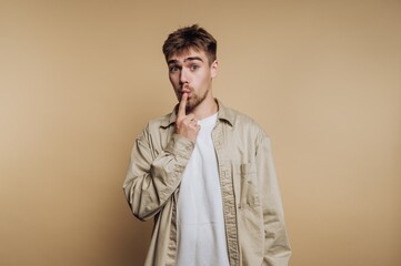 Young man making a shushing gesture against a plain backdrop while wearing a light shirt