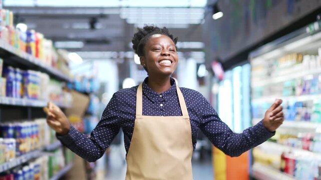 Happy cheerful african american supermarket saleswoman dancing between rows of shelves in market grocery store. Joyful funny young female seller worker in apron rejoices at workplace at marketplace
