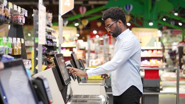 African american male shopper using a self-service cashier checkout in supermarket. Customer scanning produce items using at grocery store cash register. Man uses a terminal to pay for products online