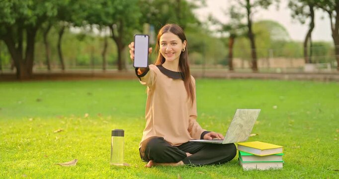 Indian young adult gen z lady hold mobile phone show white mock up blank copy space screen look camera outdoor place. Happy teen age girl female staring cam do casual pose enjoy new day at garden park