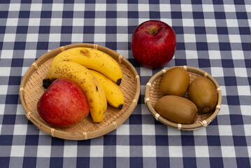 Top view of apples, bananas, and kiwis presented in two woven bamboo baskets on a navy blue and white plaid tablecloth. Ideal for healthy snack and fresh food concepts.