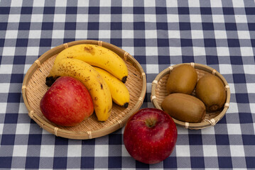 Top view of apples, bananas, and kiwis presented in two woven bamboo baskets on a navy blue and white plaid tablecloth. Ideal for healthy snack and fresh food concepts.