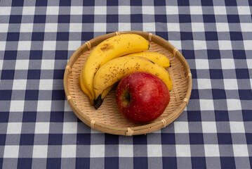 Top view of one red apple and four bananas presented in a single woven bamboo basket on a navy blue and white plaid tablecloth. Ideal for healthy snack and fresh food concepts.