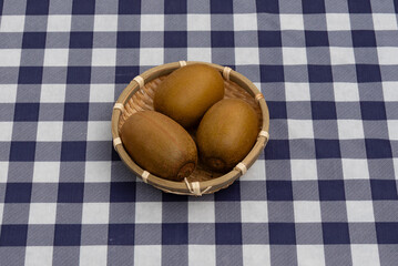 Top view of three kiwis presented in a small woven bamboo basket on a navy blue and white plaid tablecloth. Ideal for healthy snack and fresh fruit concepts.