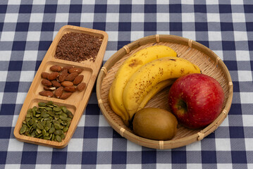 Top view of a bamboo basket with fruit (apple, bananas, kiwi) and a wooden tray with nuts and seeds (almonds, flax, pumpkin) on a navy blue plaid tablecloth. Ideal for healthy snack concepts.