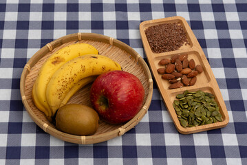 Top view of a bamboo basket with fruit (apple, bananas, kiwi) and a wooden tray with nuts and seeds (almonds, flax, pumpkin) on a navy blue plaid tablecloth. Ideal for healthy snack concepts.