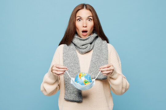Young surprised woman wear scarf sweater hold globe Earth map in sterile face mask isolated on plain blue background studio portrait. Healthy lifestyle ill sick disease treatment cold season concept.