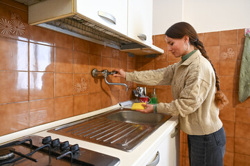 Woman in beige shirt washes dishes under running water in a traditional brown-tiled kitchen, showing everyday life and home responsibility.
