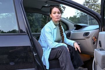 Confident woman sits in the doorway of her car, dressed casually with a braid, ready to travel or take a short break on a scenic Italian road.
