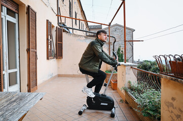 Focused young man in a green jacket squats outdoors on a terrace, practicing balance and strength exercises during a calm Italian morning.
