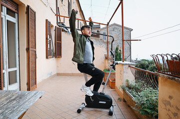 Active young man in a green jacket performs squats on a terrace, focusing on fitness and a healthy lifestyle in an open-air Italian setting.
