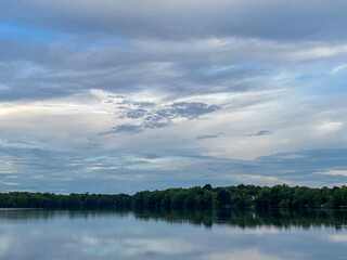 Symmetrical landscape of a calm lake with a perfect mirror reflection of the cloudy sky and a dense green forest treeline.