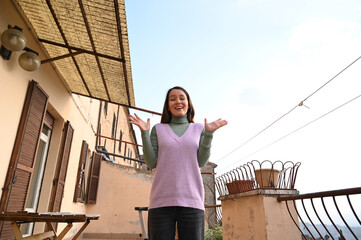 Joyful young woman in a purple dress raises her hands with excitement under bright sunlight on a balcony in a Mediterranean town.
