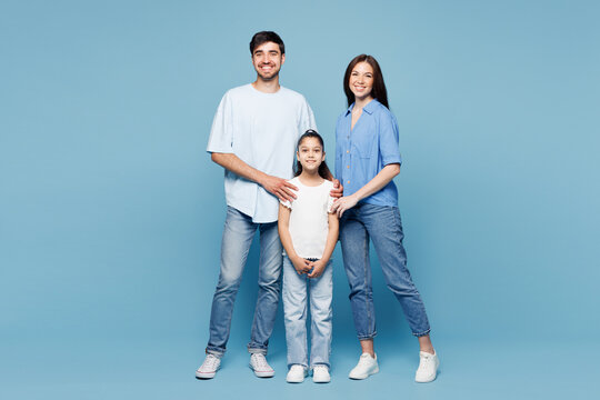 Full body young smiling lovely cheerful happy parents mom dad with child kid daughter girl wearing casual clothes stand hug look camera isolated on plain blue background. Family day childhood concept.
