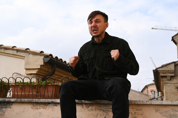 A young man in a black jacket sitting outdoors shows intense emotions — shouting passionately with clenched fists, expressing motivation and determination.
