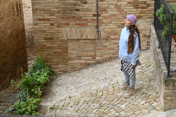 Young woman in blue hoodie gazes upward near an old rustic brick wall in sunlight, expressing curiosity and peace in an Italian street scene.