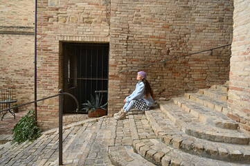 Elderly man sits and rests on ancient stairs surrounded by weathered brick walls, enjoying a calm moment in a picturesque Italian village.
