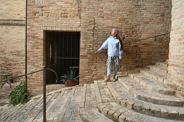 Stylish elderly man in casual blue jacket posing on narrow cobblestone street between rustic brick walls, symbolizing freedom, travel, and Italian charm.
