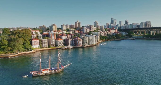 Australia, Sydney: old fashioned tourist ship sailing Sydney Harbor water, residential buildings lining the shore and the city skyline with skyscrapers and a bridge in the background under a blue sky