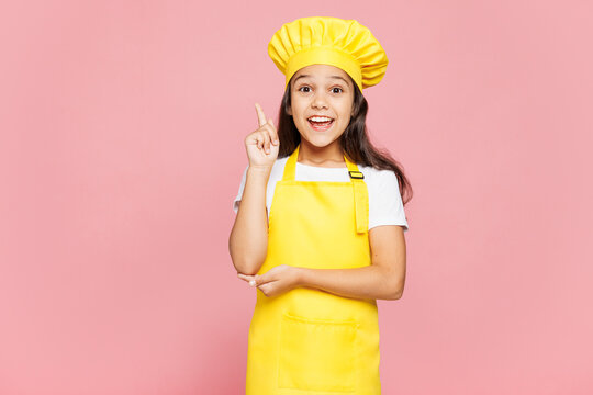 Small kid child girl housewife chef cook baker mother's helper wear yellow apron toque cap holding index finger up with great new idea isolated on pastel plain pink background. Cooking food concept.