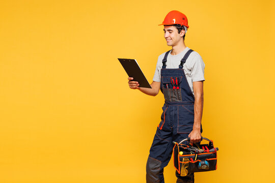 Young employee laborer handyman man wears helmet overall hold clipboard paper account documents toolbox isolated on plain yellow background. Instruments renovation apartment room. Repair home concept.