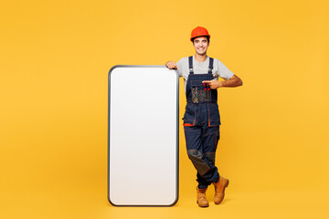 Full body young employee laborer handyman man wear helmet overall point finger on big huge blank screen mobile cell phone isolated on plain yellow background Instruments for renovation apartment room