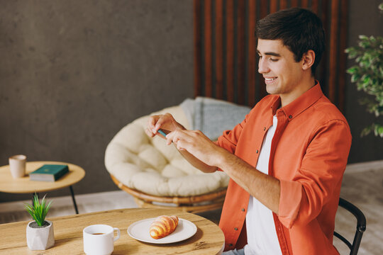 Young man wear casual clothes taking cell phone picture of croissant sitting alone at table in coffee shop cafe relax rest in restaurant in free time indoors. Freelance mobile office business concept.